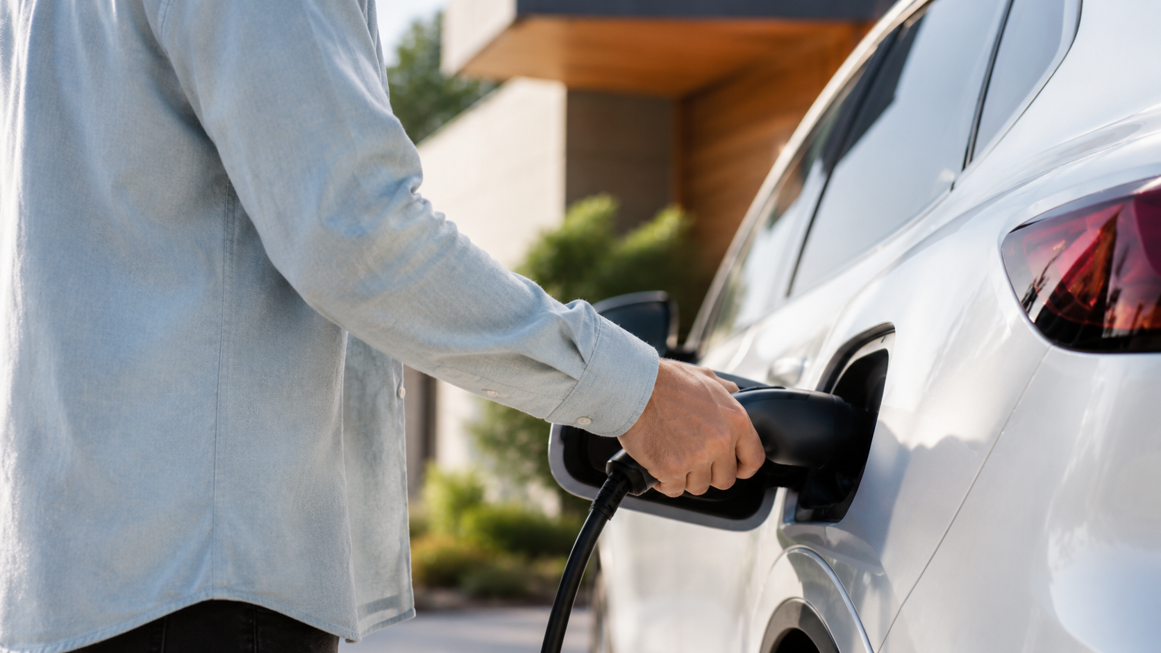 Person in a light denim shirt plugs a charging cable into a white electric car outside.